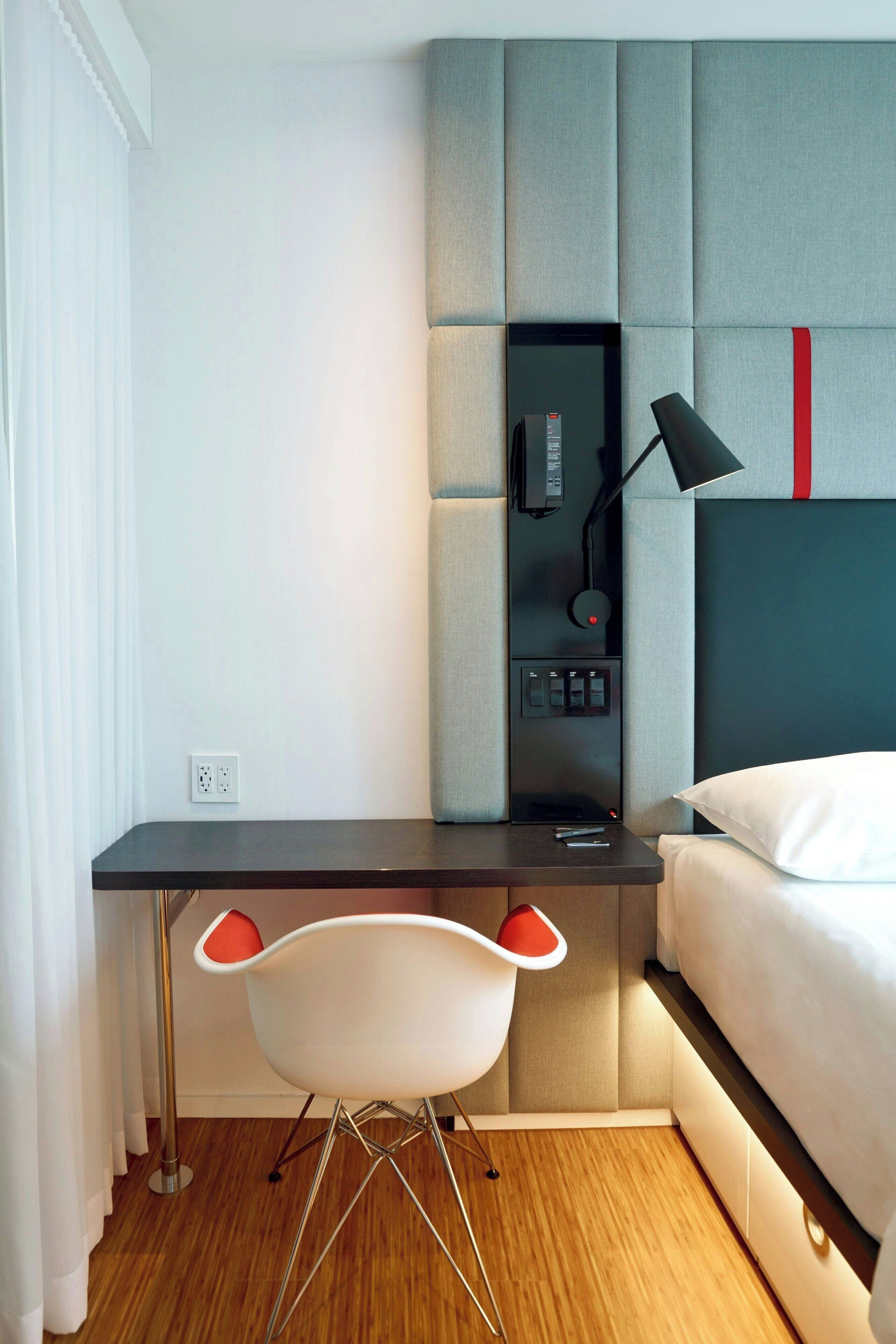 A modern bedroom with a bed, a desk with a chair, a bedside table, a lamp, and a power outlet.