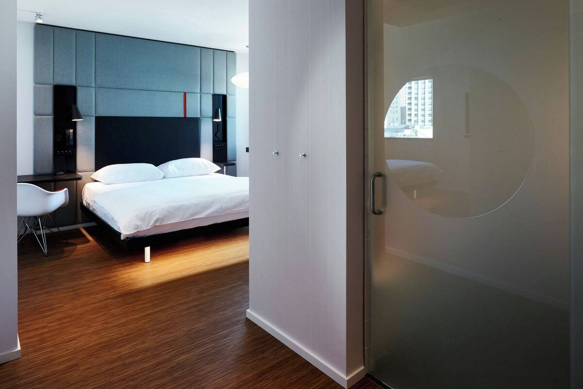 A modern hotel room with a bed, a desk, and a chair. There is a wardrobe and a window with a view of buildings.