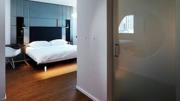 A modern hotel room with a bed, a desk, and a chair. There is a wardrobe and a window with a view of buildings.