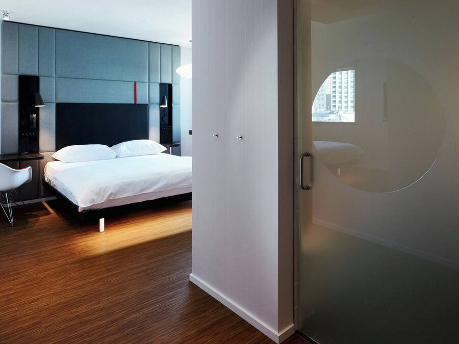 A modern hotel room with a bed, a desk, and a chair. There is a wardrobe and a window with a view of buildings.