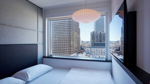 Ett modernt hotellrum med ett stort fönster som erbjuder utsikt över staden, en snyggt bäddad säng och en hängande lampa.
