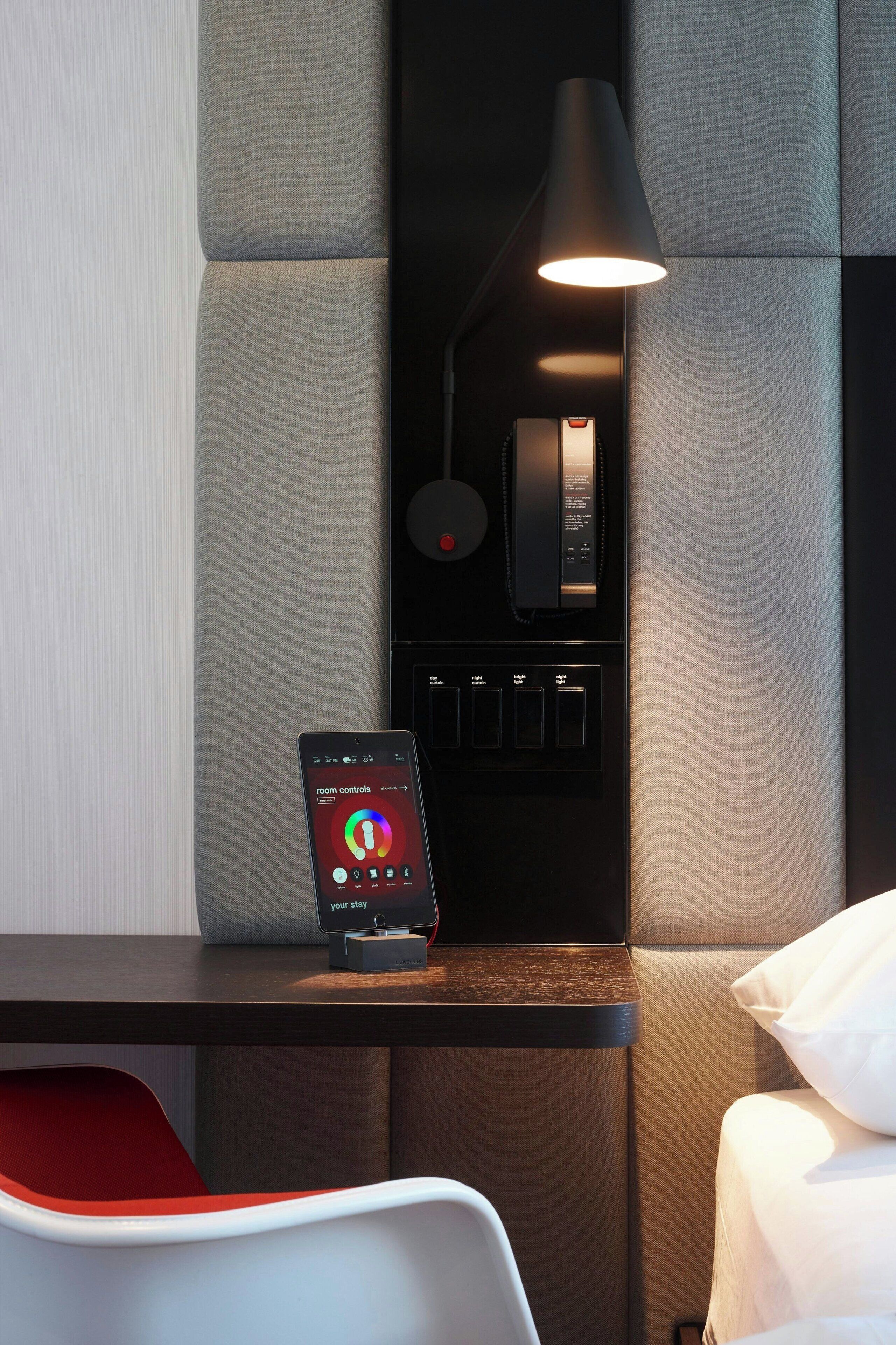 Ein modernes Hotelzimmer mit Bedienfeld, einem Smartphone, das Zimmerfunktionen anzeigt, und einem Bett mit weißer Bettwäsche.