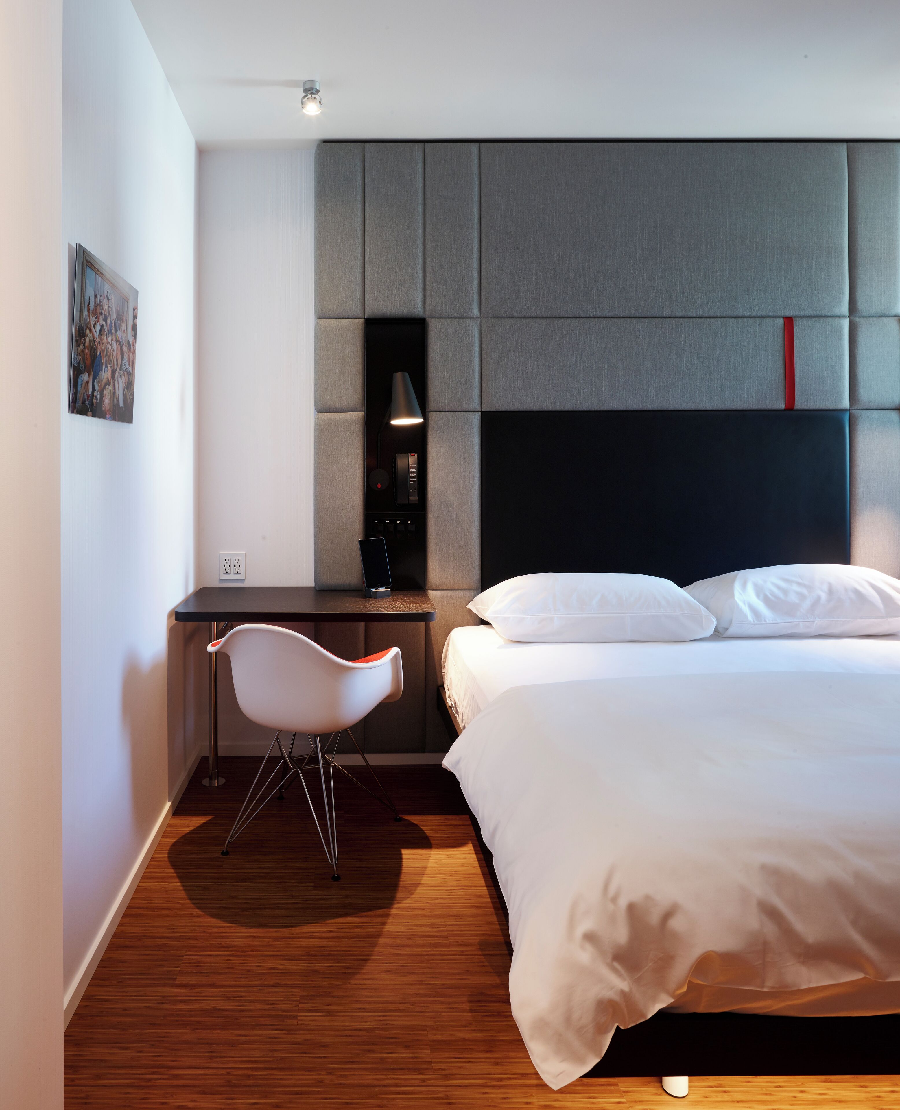 A modern hotel room with a wooden floor, a bed with white linens, a desk with a chair, and a headboard with a red accent.