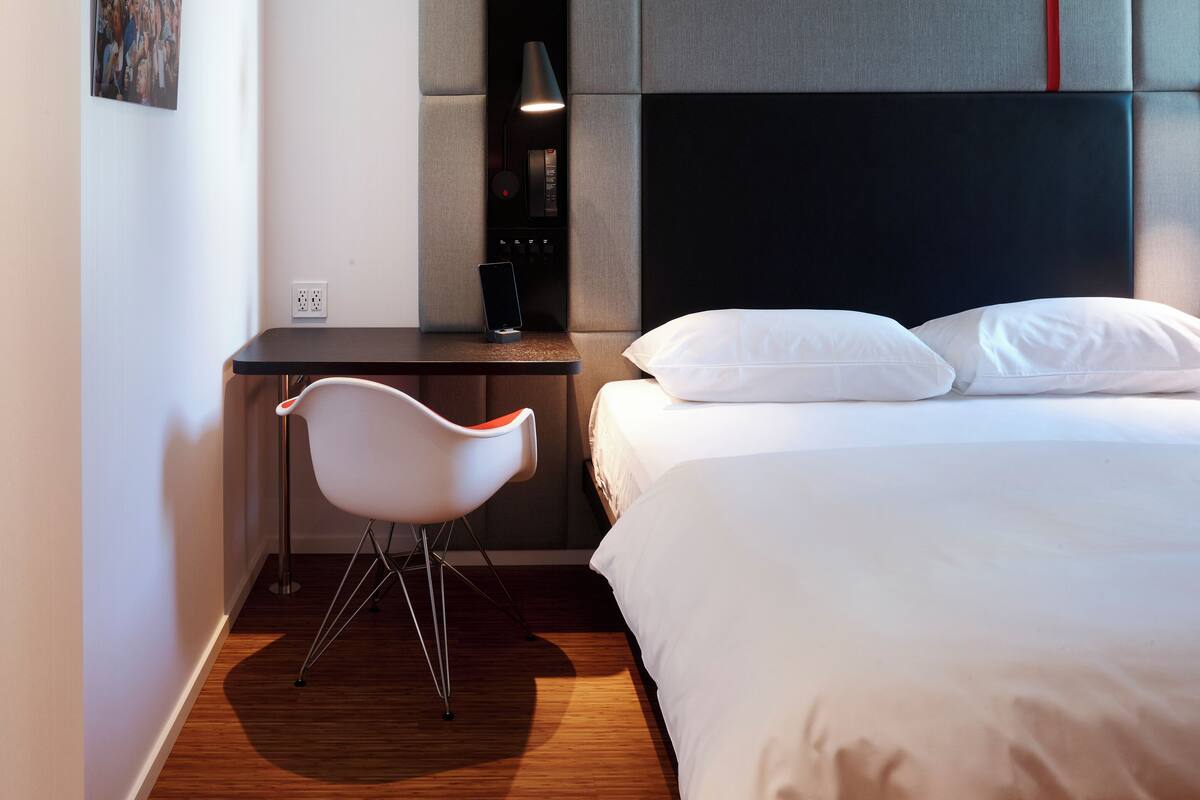 A modern hotel room with a wooden floor, a bed with white linens, a desk with a chair, and a headboard with a red accent.