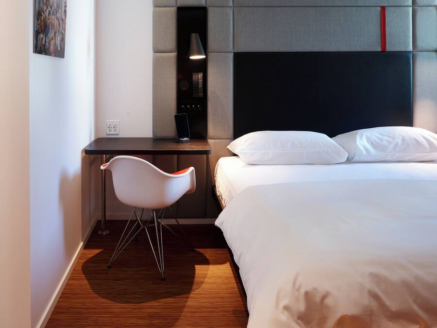 A modern hotel room with a wooden floor, a bed with white linens, a desk with a chair, and a headboard with a red accent.
