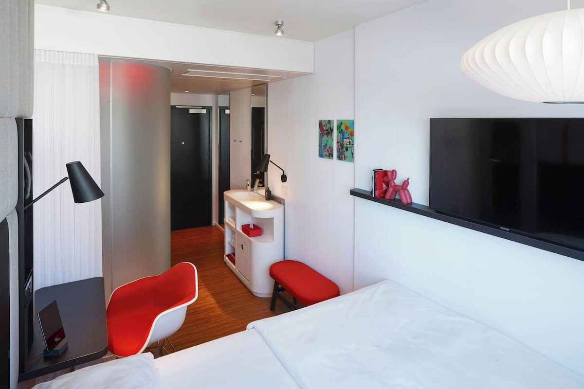 A modern hotel room with a bed, a red chair, a desk with a lamp, a TV, and a small bathroom.