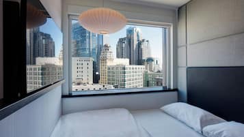 A hotel room with a large window offering a city view, a bed with white linens, and a modern pendant light.