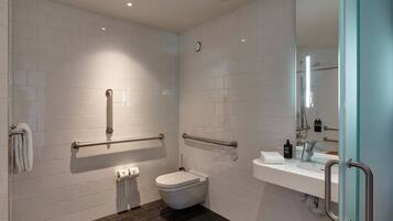 A modern bathroom with white tiled walls, a dark floor, a toilet, a sink with a mirror, and handrails.