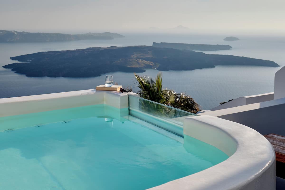 A clear blue sky with a view of a calm sea and a white infinity pool with a glass of water and a book on the edge.