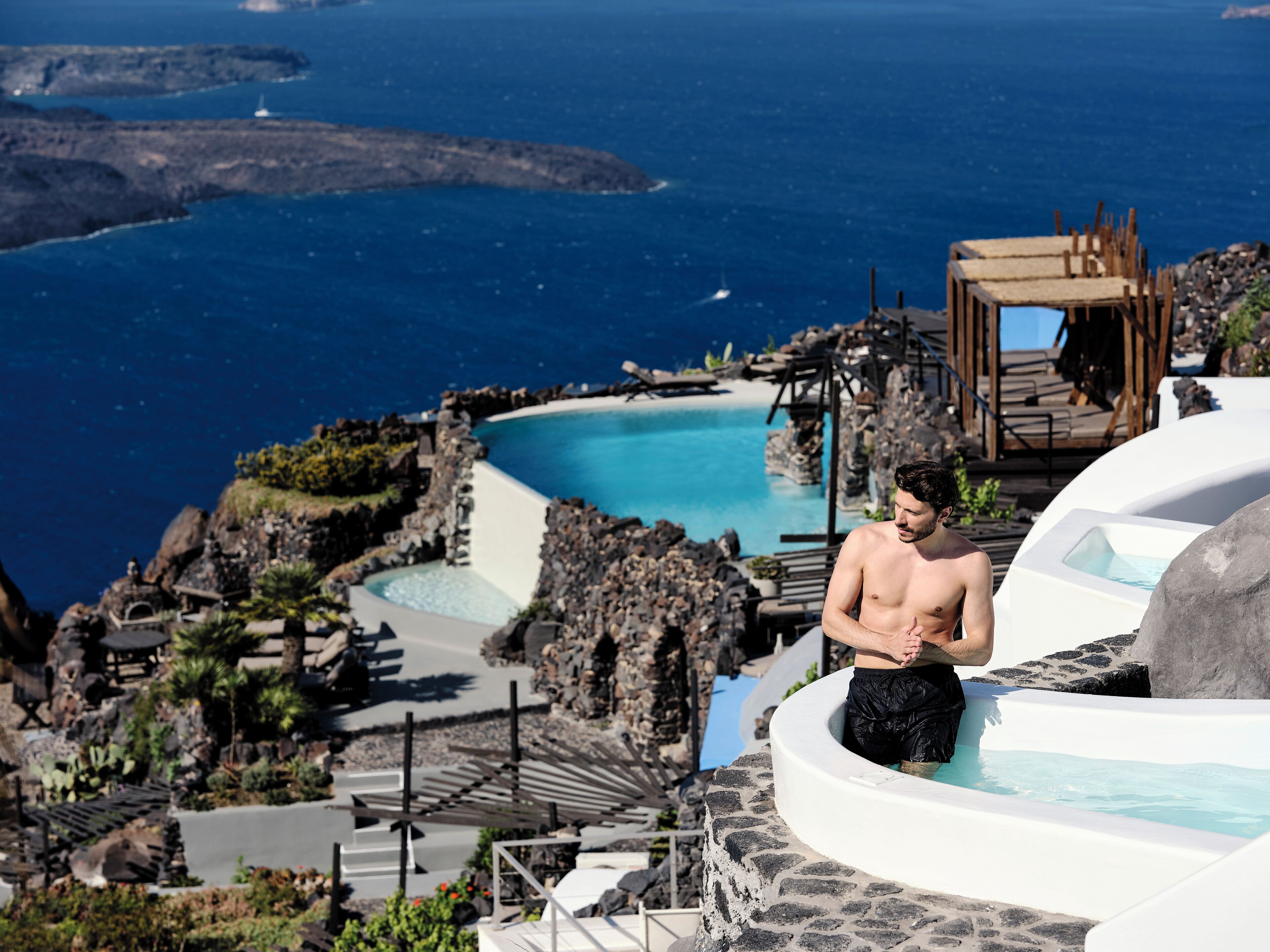 Un hombre en un jacuzzi con vista a una piscina y al mar, con terreno rocoso al fondo.