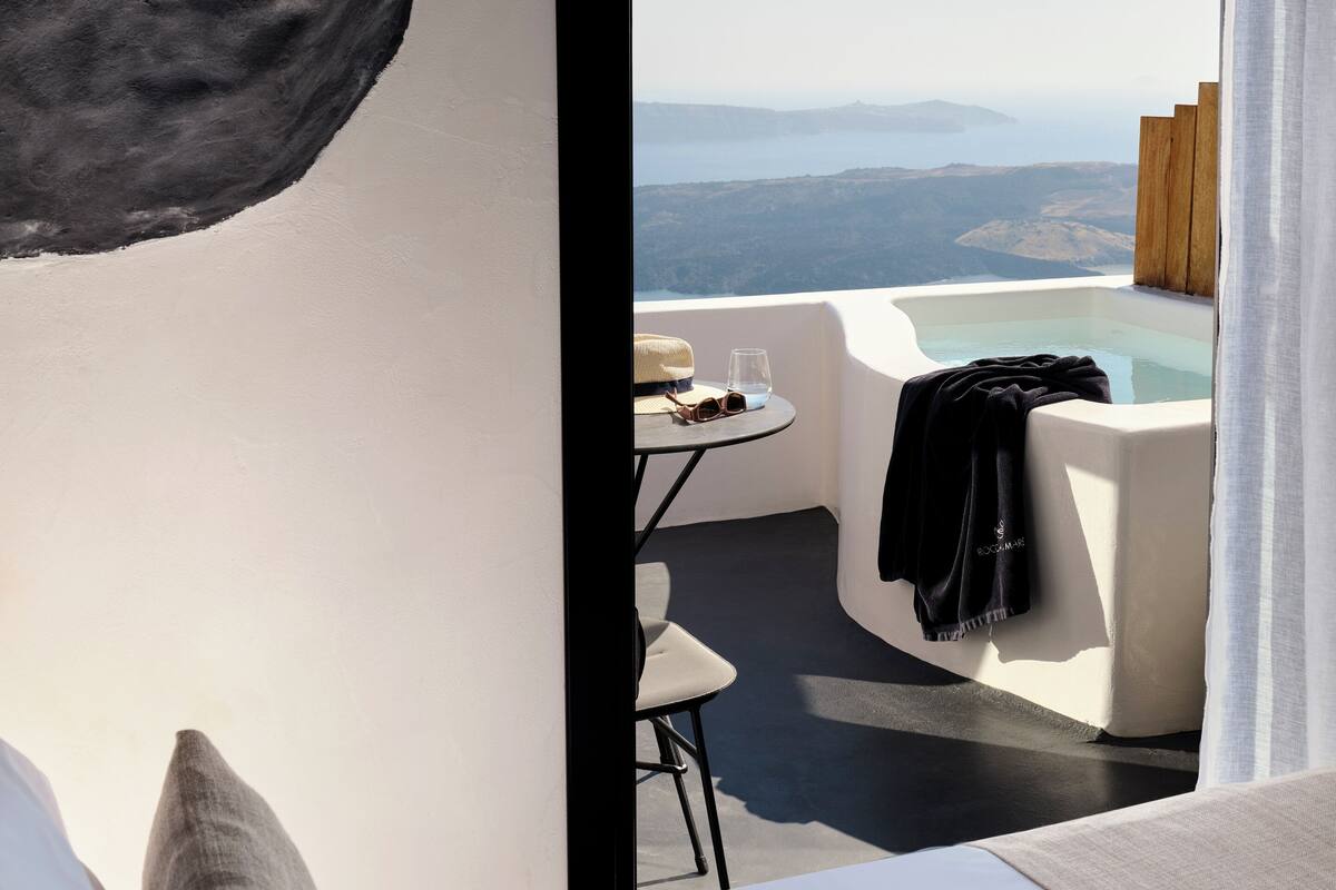 A modern bedroom with a large bed, a small table with a glass of water and sunglasses, and a view of a mountain landscape through a large window.