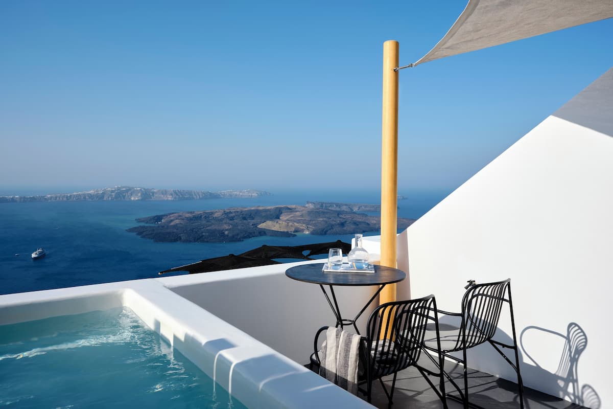 A balcony with a view of the sea, a small pool, a table with chairs, and a bottle.