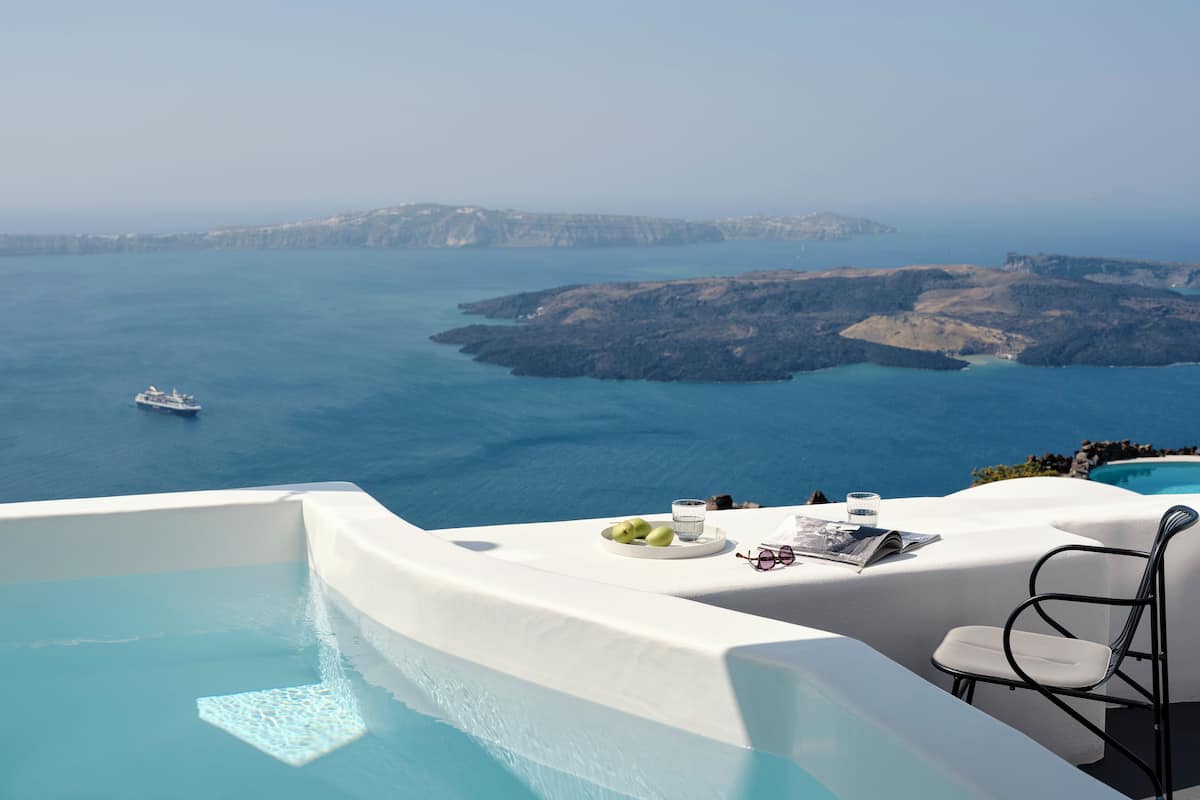 A poolside area with a view of the sea and cliffs, a boat in the distance, and a table with glasses and a plate of fruit.
