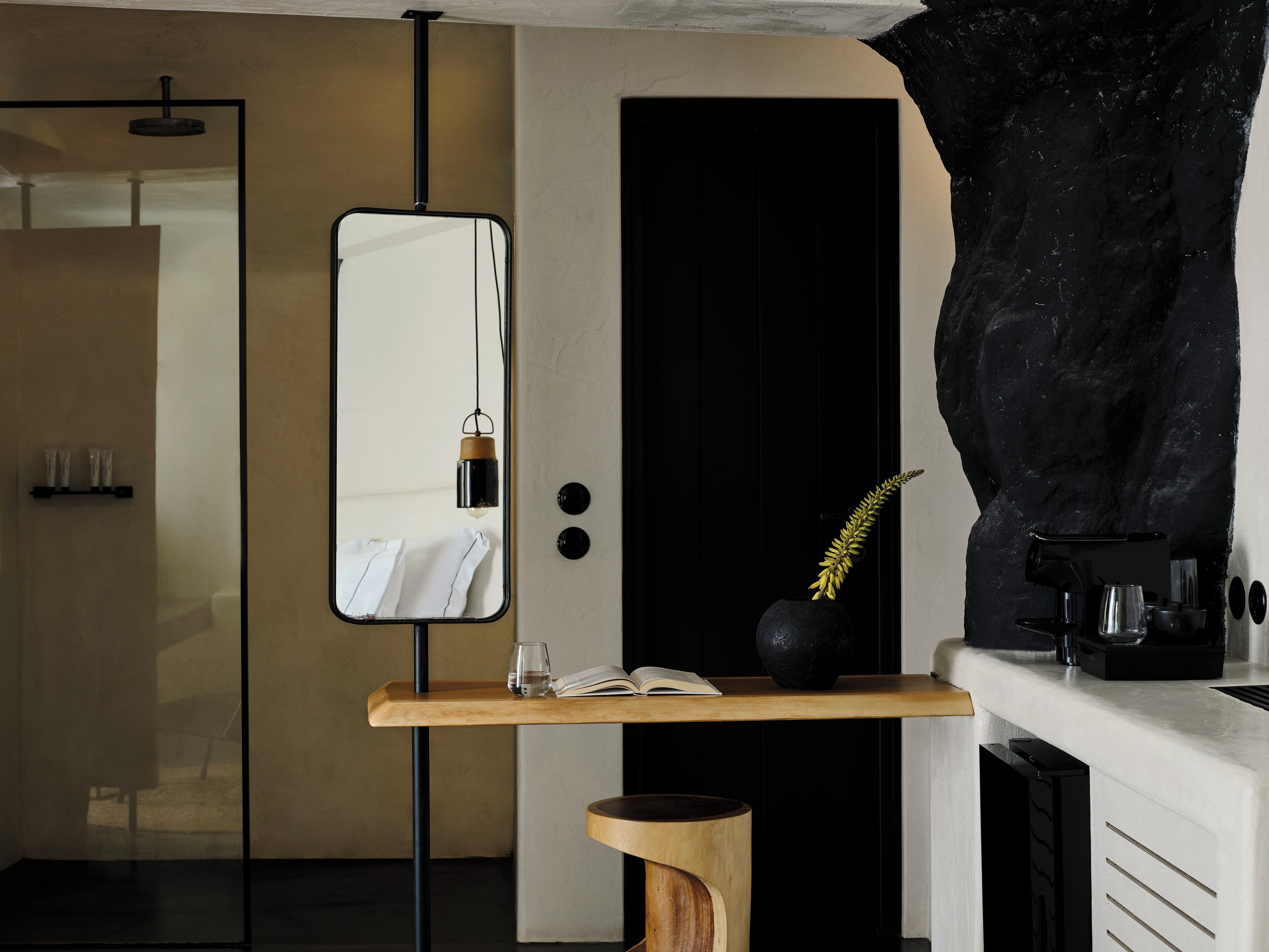 Un baño moderno con espejo, un taburete de madera, un florero negro con planta y una cafetera.