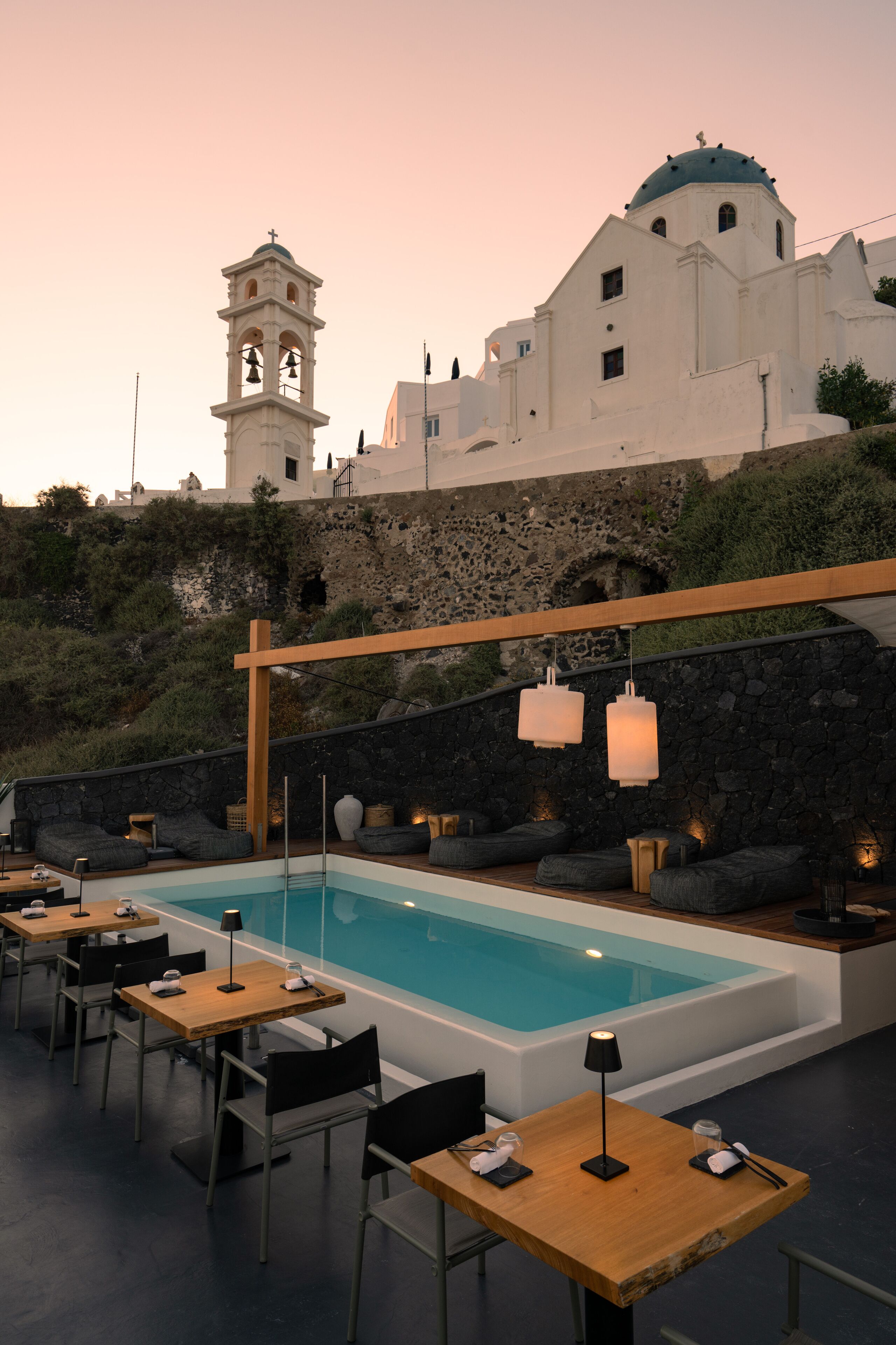 A poolside area with tables and chairs, a stone wall, and a bell tower in the background.