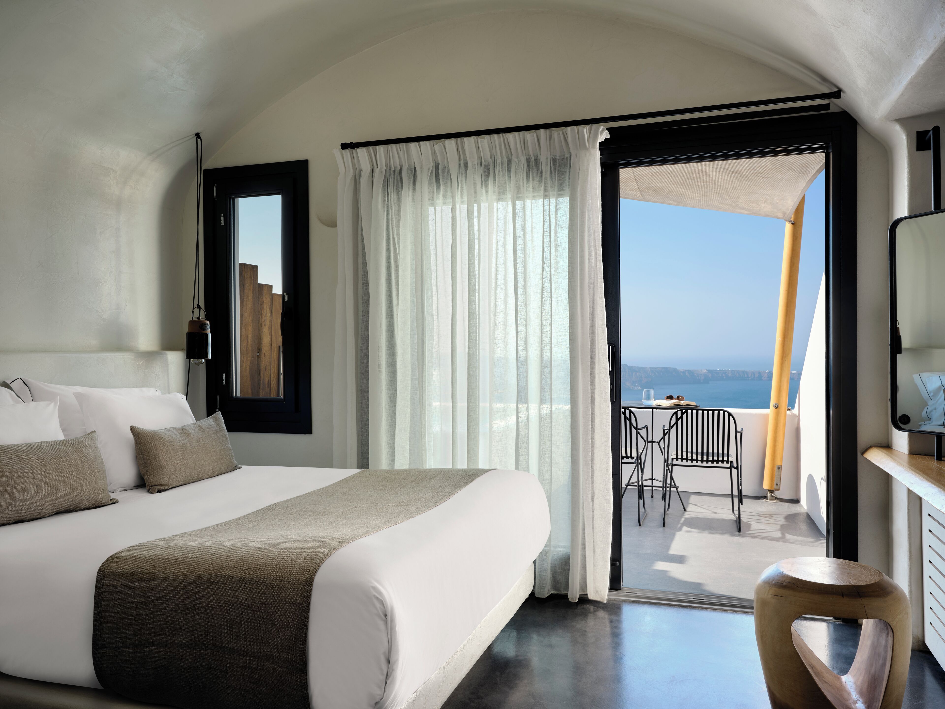 A modern hotel room with a large bed, a balcony with a table and chairs, and a view of the sea.