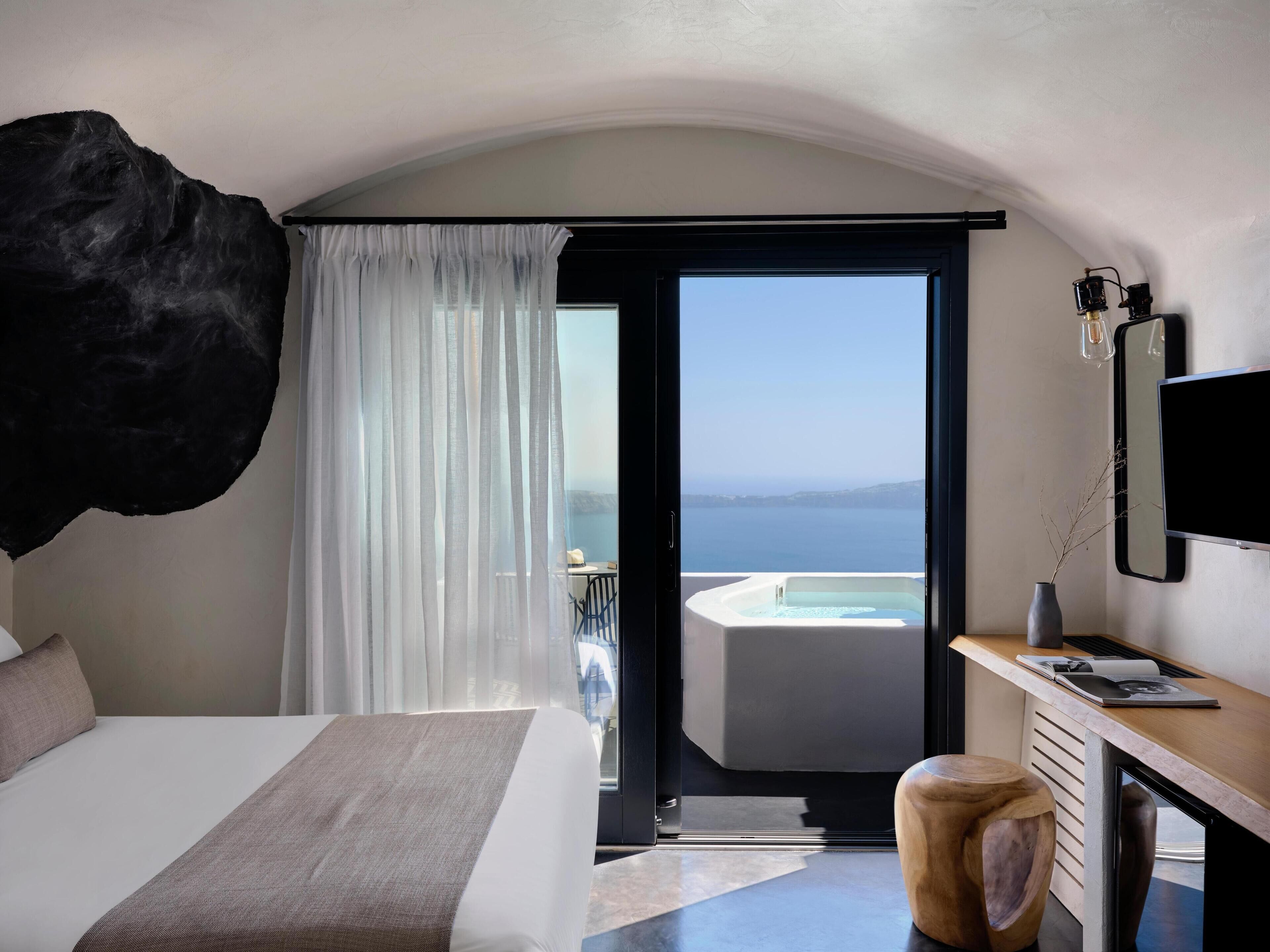 Habitación de hotel moderna con una cama grande, un escritorio con computadora, vista al mar y un jacuzzi.