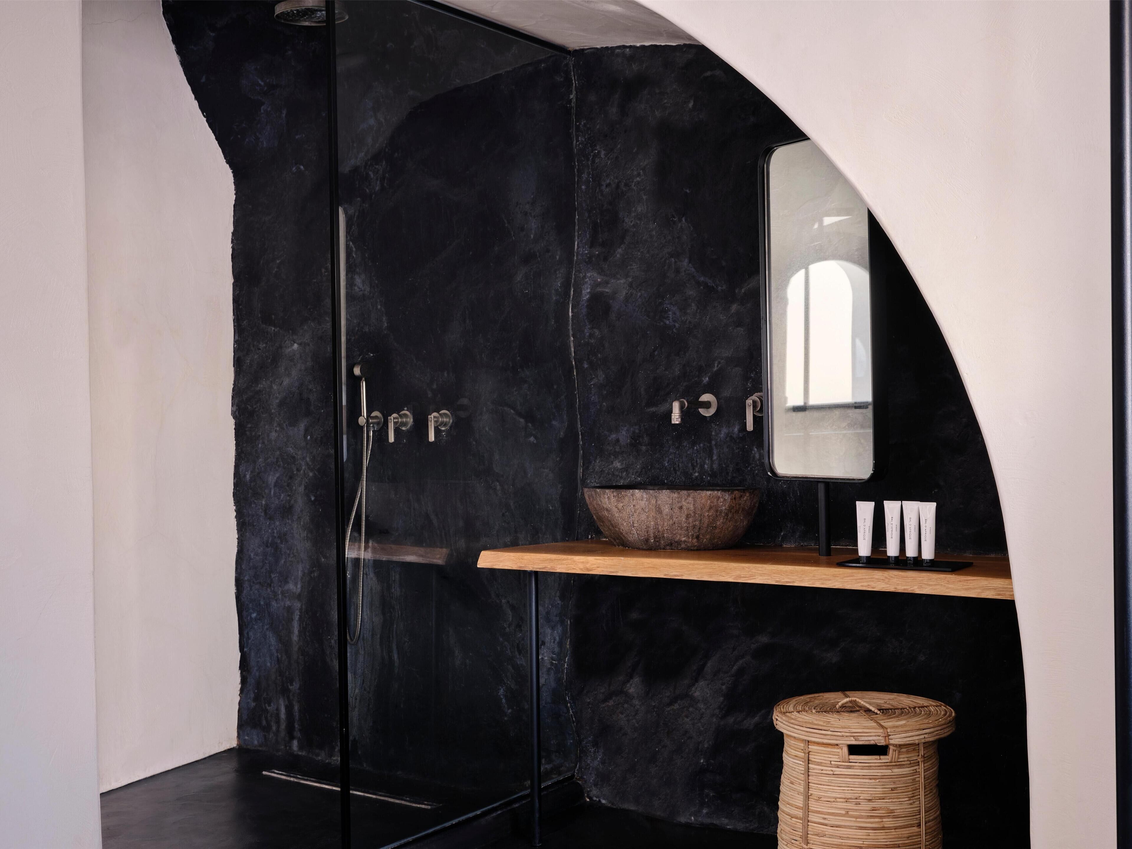 A modern bathroom with a black marble wall, a glass shower enclosure, a wooden shelf with toiletries, and a woven basket.