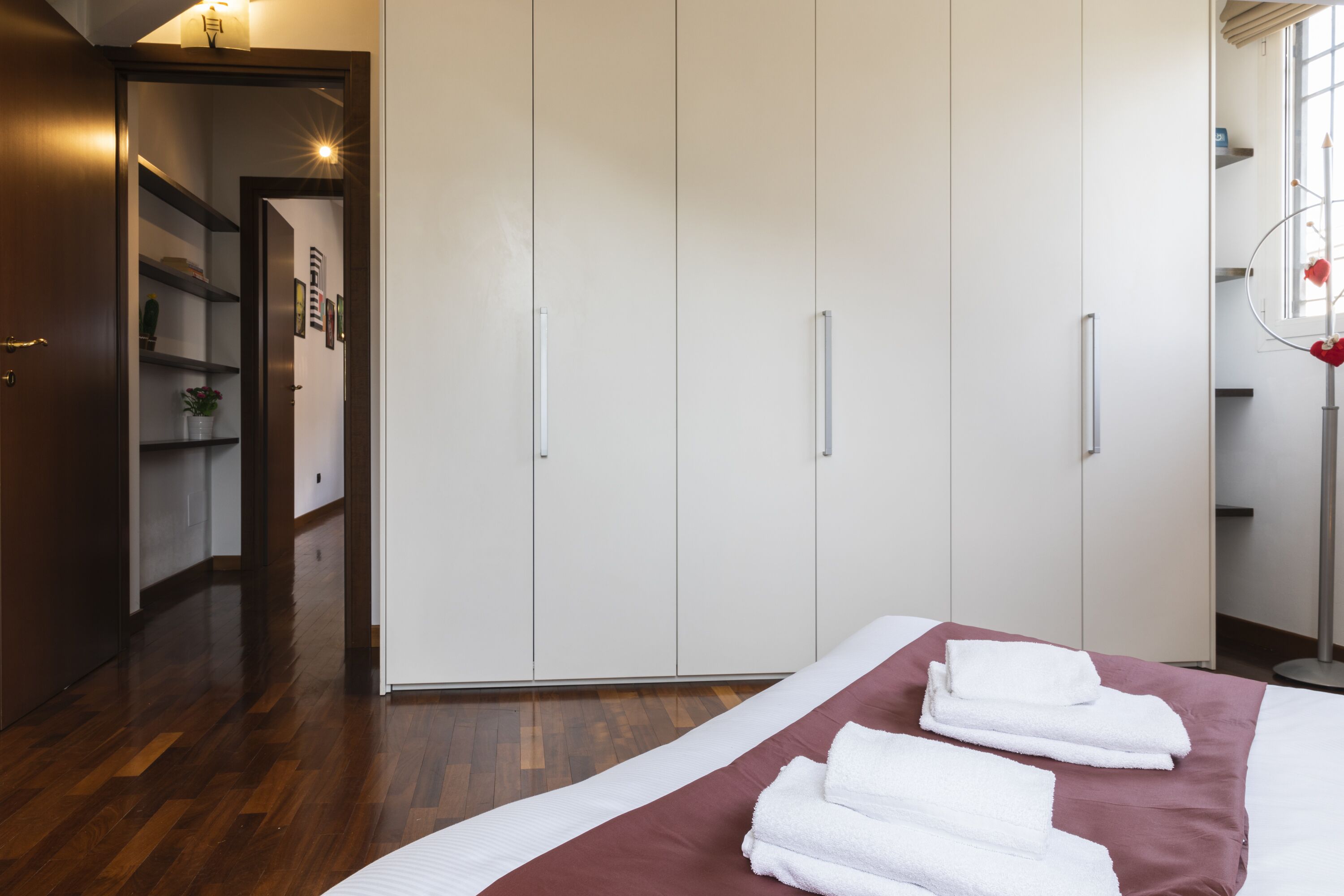 A hotel room with a large wardrobe, a bed with a red and white bedspread, and a wooden floor.