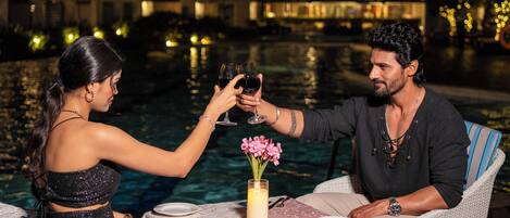 Una pareja brindando con copas de vino en una mesa junto a una piscina, con una vela y flores sobre la mesa.