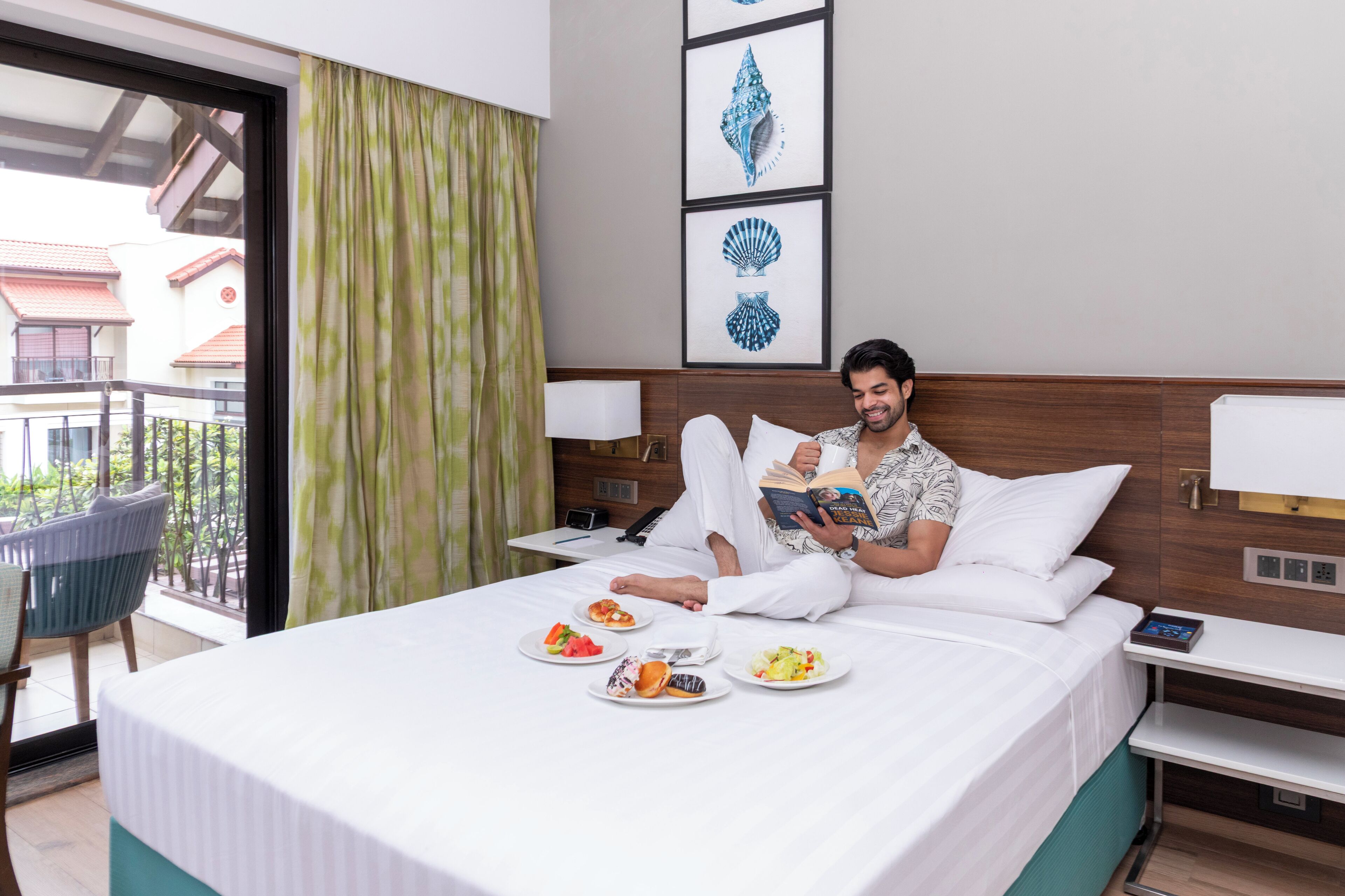 A hotel room with a bed, a bedside table with a lamp, a chair, and a balcony with a view of buildings and trees.