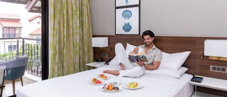 A hotel room with a bed, a bedside table with a lamp, a chair, and a balcony with a view of buildings and trees.