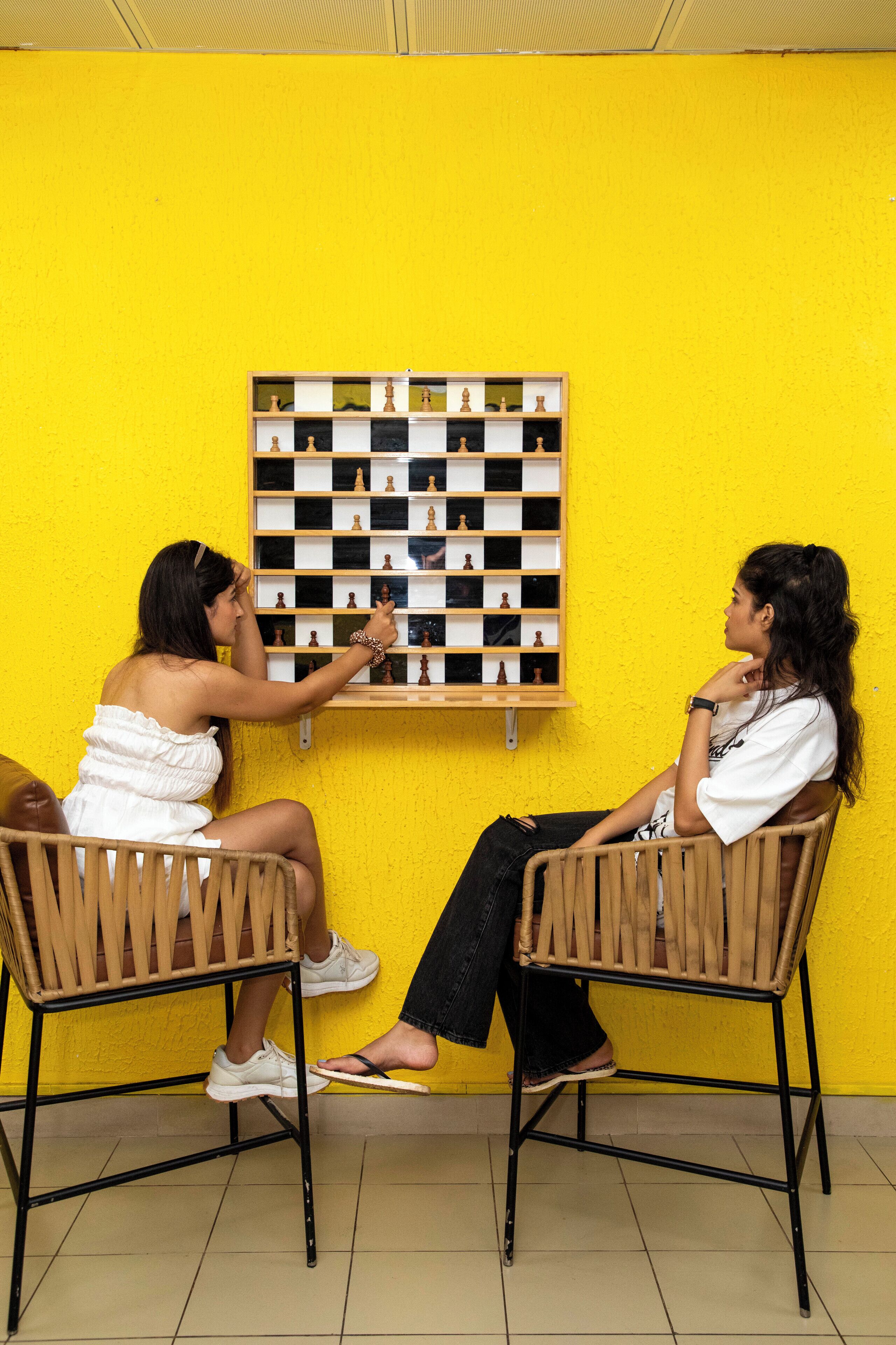 Two individuals seated on wicker chairs facing a chessboard against a yellow wall.