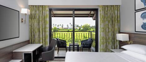 Habitación de hotel con cama, escritorio y sillas, un balcón con vistas a la vegetación y un televisor montado en la pared.