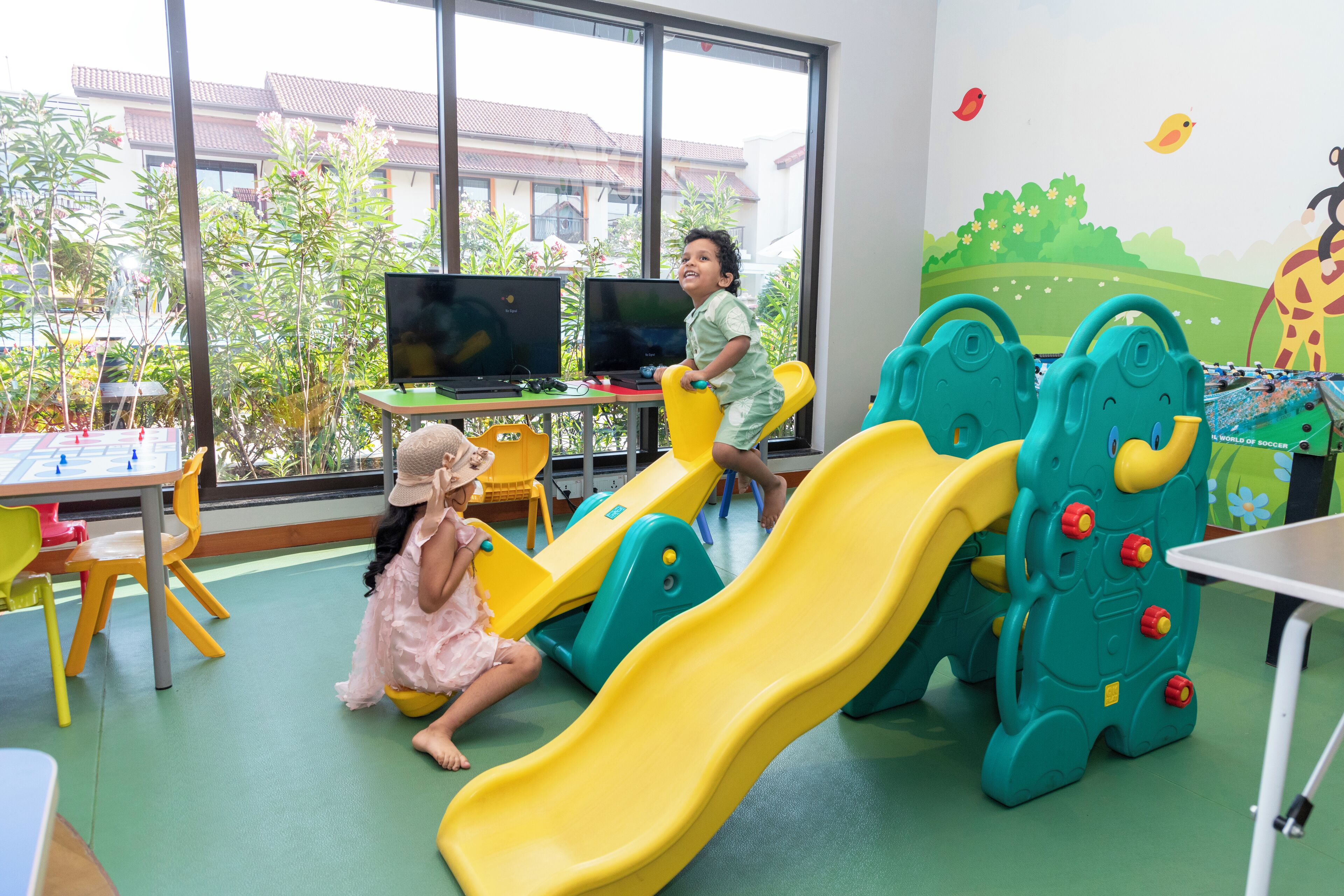 A children's play area with slides and climbing structures, a table with a laptop, and a view of a building through large windows.