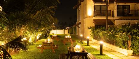 Un espacio al aire libre bien iluminado con mesas de picnic y luces colgantes, rodeado de abundante vegetación y un edificio con balcones.