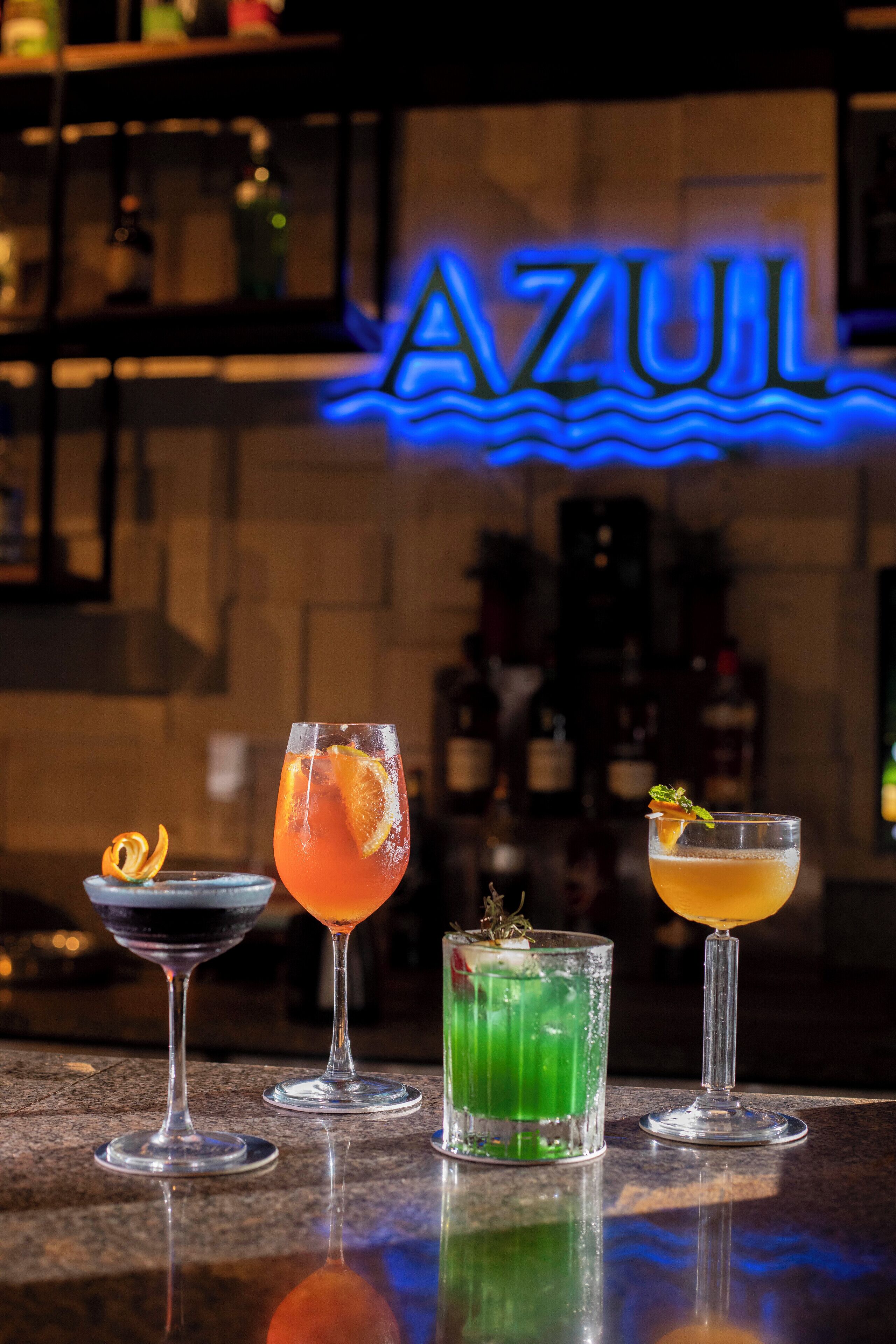 A bar area with four different cocktails on the counter, a neon sign reading "AZUL" in the background, and a reflection on the polished surface.