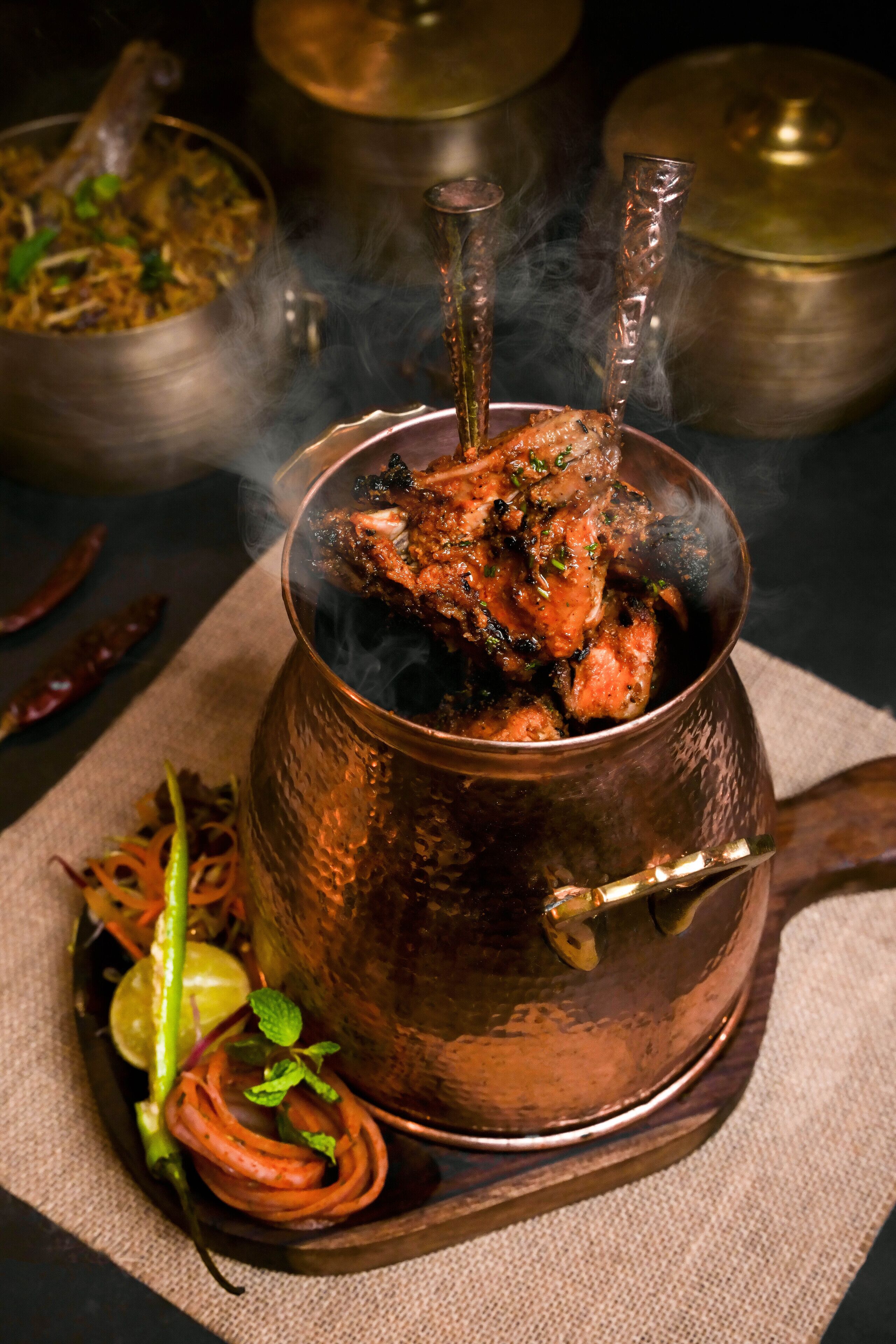 A copper pot with a lid, containing a dish with a piece of meat and herbs, surrounded by a cloth and some vegetables.