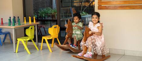 Two children on rocking chairs in a room with a tiled floor, a table with chairs, and a wall with photos.