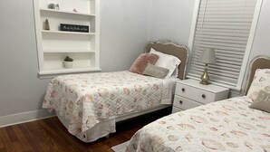 A bedroom with two beds, a nightstand, a shelf with decorative items, and a window with blinds.