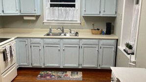 A kitchen with light blue cabinets, a wooden floor, a window with a white curtain, a sink, and a coffee maker.