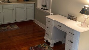 A kitchen with white cabinets, a white countertop, a window with a white curtain, a painting on the wall, and a white desk with drawers.
