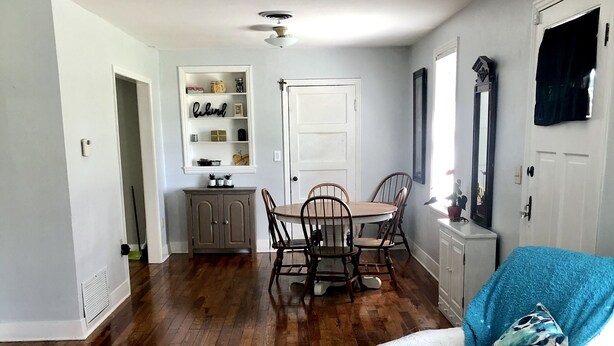 A spacious living room with hardwood floors, a dining area with a wooden table and chairs, a white sofa with a blue throw, and a white door.