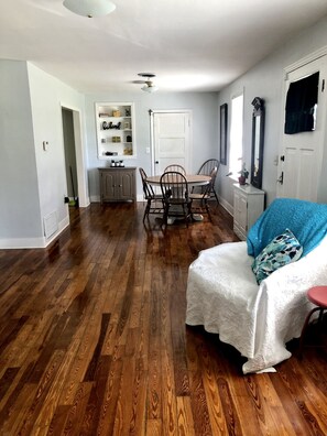 Un salon spacieux doté de planchers en bois, un coin repas avec une table et des chaises en bois, un canapé blanc agrémenté d’un plaid bleu et une porte blanche.