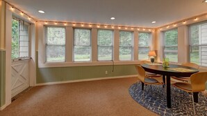 A dining area with a round table, four chairs, a patterned rug, and string lights along the windows. - Sophisticated Lenox Retreat by Evergreen Home (Lenox)