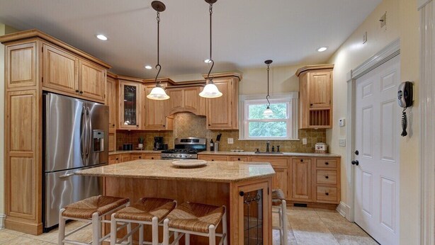 A kitchen with wooden cabinets, a stainless steel refrigerator, a central island with bar stools, pendant lighting, and a window providing natural light. - Sophisticated Lenox Retreat by Evergreen Home (Lenox)
