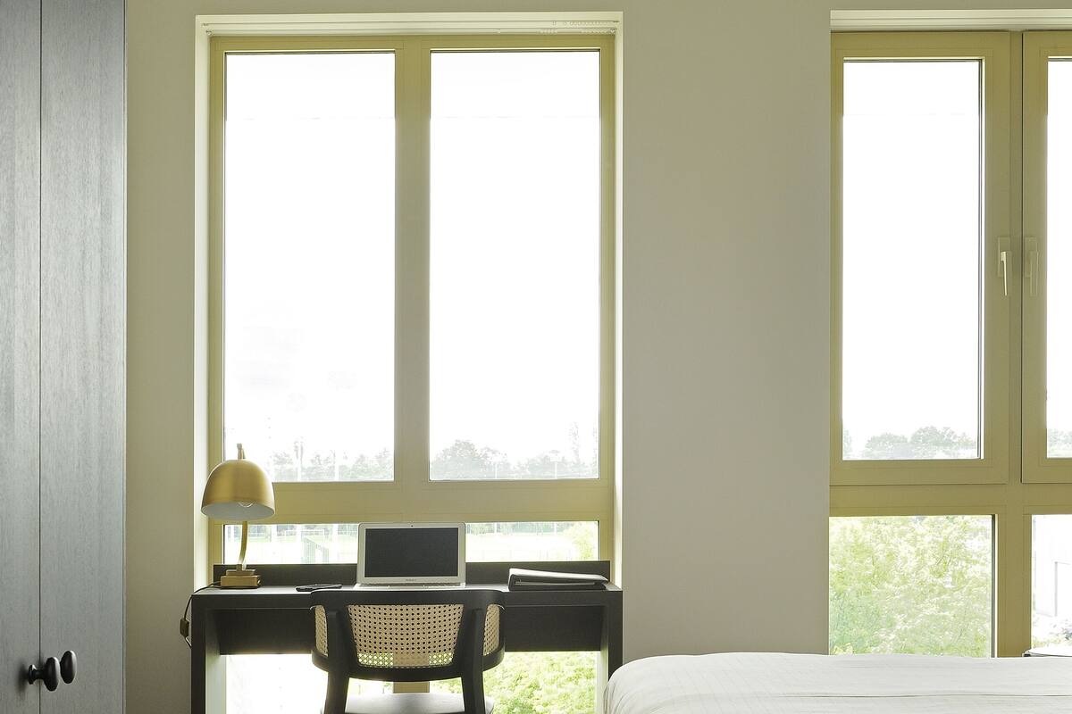 A bedroom with a bed, a desk with a chair, and large windows.