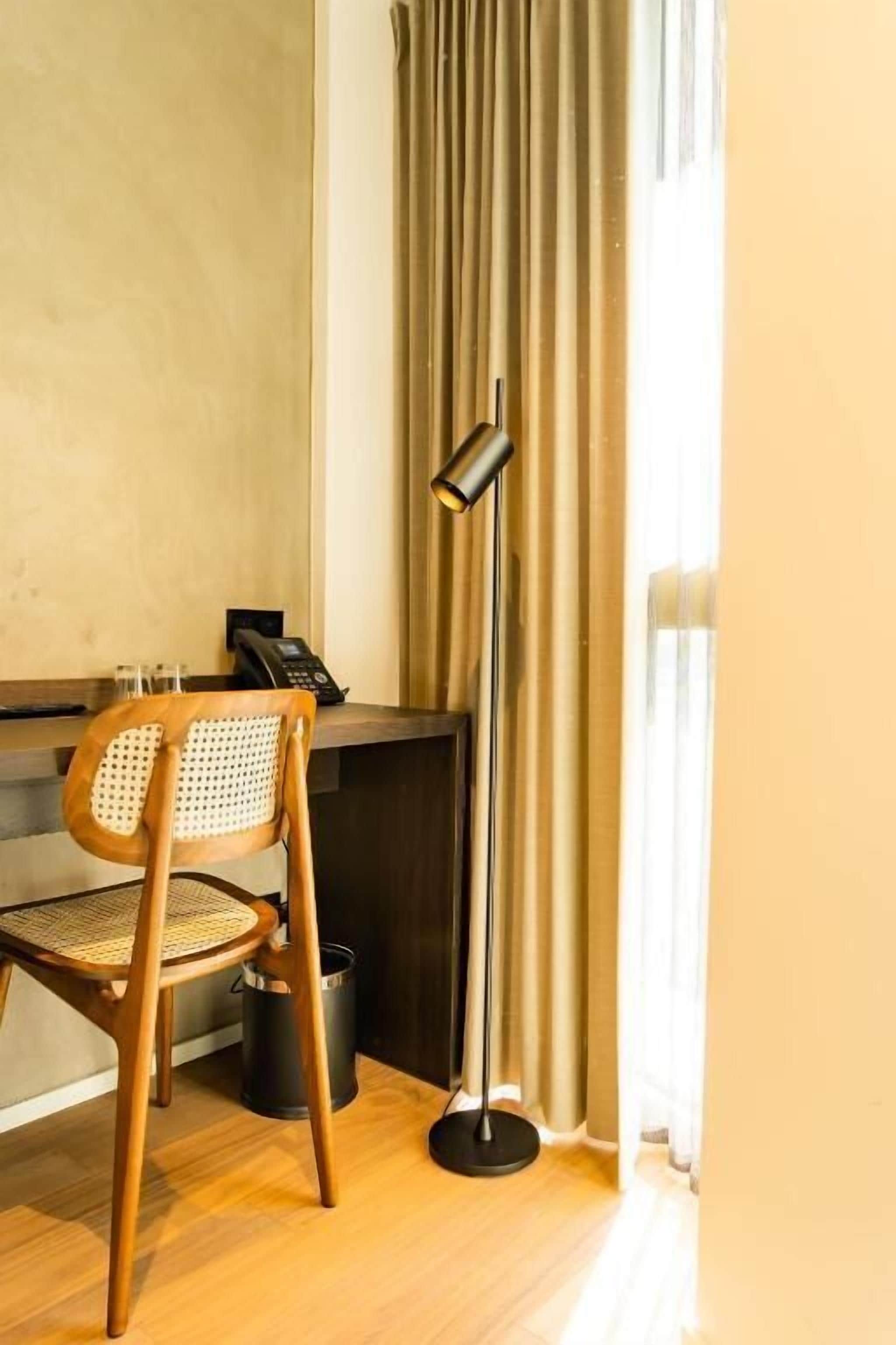 A hotel room with a wooden chair, a desk with a telephone, a floor lamp, and a window with curtains.