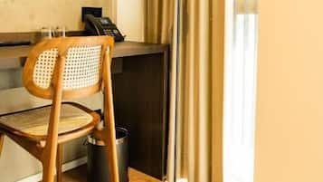 A hotel room with a wooden chair, a desk with a telephone, a floor lamp, and a window with curtains.