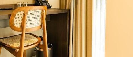 A hotel room with a wooden chair, a desk with a telephone, a floor lamp, and a window with curtains.