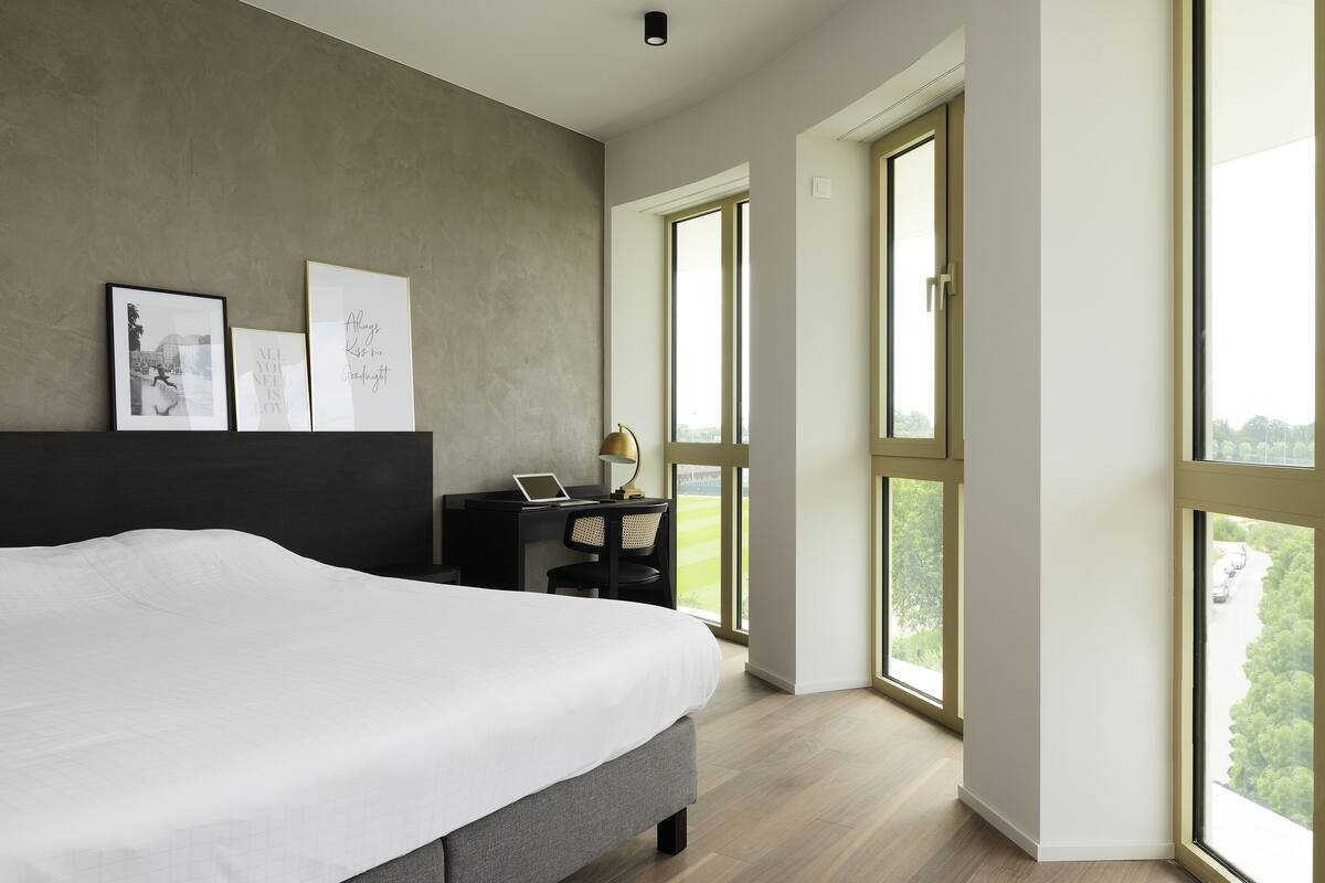 A modern bedroom with a large bed, a desk with a laptop, and large windows offering a view of the outdoors.