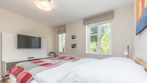 A modern bedroom with a large bed, a flat-screen TV mounted on the wall, and a window with a view of greenery.