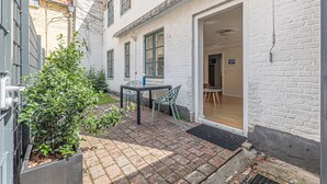 A courtyard with a table and chairs, a potted plant, and a building with a white brick exterior and a wooden door.