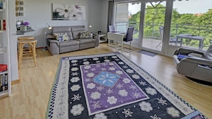 A modern living room with a grey sofa, a small desk, a large window with a balcony, and a patterned rug.
