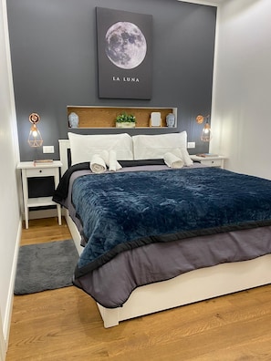Quarto com uma cama grande, mesa de cabeceira, luminária na parede e um quadro com a lua.