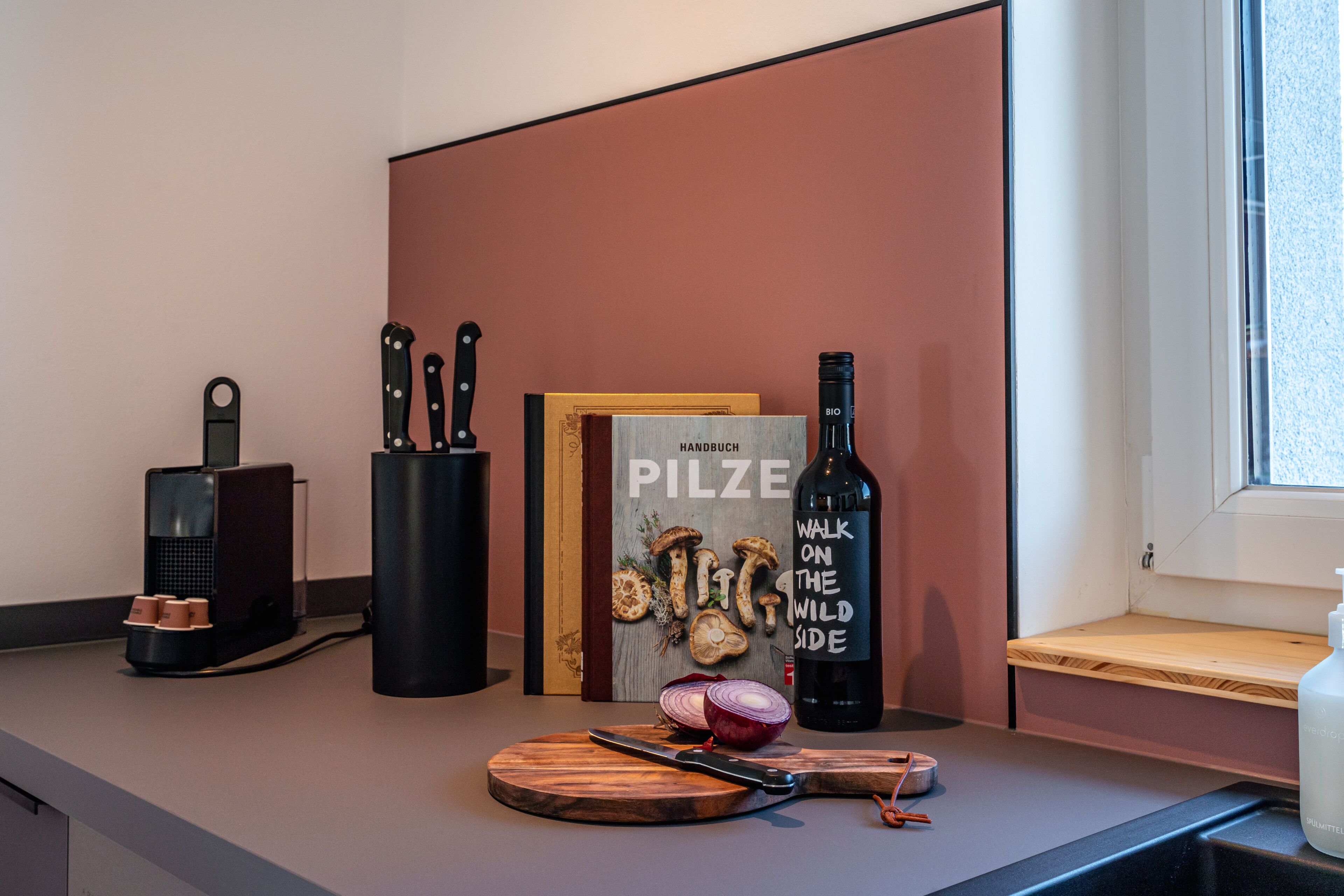 Una encimera de cocina con tabla de cortar, un cuchillo, una botella de vino, una máquina de café y un libro titulado 'PILZE'.